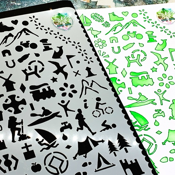 Adventures Journal Stencil, Travel A5 Journal Sized Stencil with adventure activity shapes & OS Symbols, craft mylar stencil - showing a close up of the right hand side with train, bird, OS map symbols, mountains, cyclist, runner, tap, swimmer, information. Just off the courner of the stencil you can see a journal page poking out beneath where you can see the stencil has been used with green inks and pens.