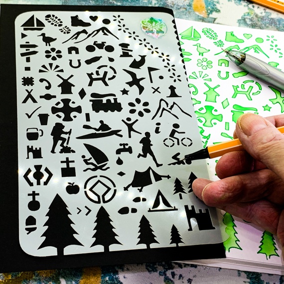Adventures Journal Stencil, Travel A5 Journal Sized Stencil with adventure activity shapes & OS Symbols, craft mylar stencil showing activity shapes with a hand appearing in bottom right hand corner holding a fine line pen about to use the stencil, behind the stencil you can see the edge of a journal where the stencil has been used with green inks