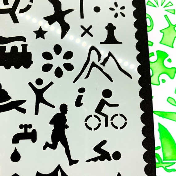 Adventures Journal Stencil, Travel A5 Journal Sized Stencil with adventure activity shapes & OS Symbols, craft mylar stencil - showing a close up of the right hand side with train, bird, OS map symbols, mountains, cyclist, runner, tap, swimmer, information. Just off the courner of the stencil you can see a journal page poking out beneath where you can see the stencil has been used with green inks and pens.