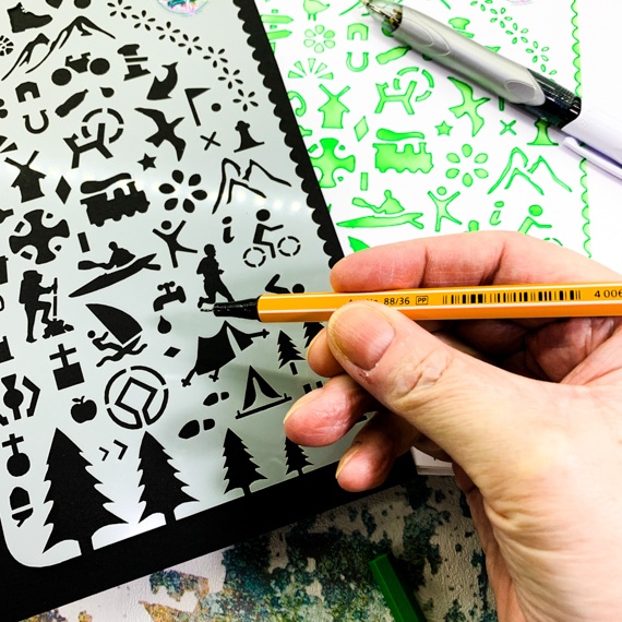 Adventures Journal Stencil, Travel A5 Journal Sized Stencil with adventure activity shapes & OS Symbols, craft mylar stencil showing activity shapes with a hand appearing in bottom right hand corner holding a fine line pen about to use the stencil, behind the stencil you can see the edge of a journal where the stencil has been used with green inks