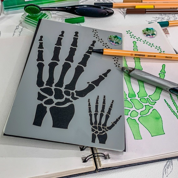 Anatomical skeleton hand stencil with a mini version along side laying on an open journal overlaying a page where the stencil has been used with green ink, with an open pen and propelling pencil laying across the journal and stencil.