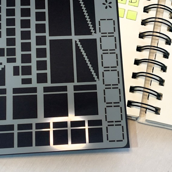 image shows close up of mylar stencil for layout basics showing boxes, squares, oblongs and dividers. poking out of the stencil to the right is the central spiral of the open journal