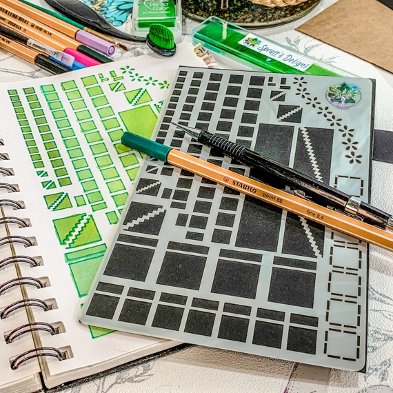 Image of the Journal Layouts Spreads Stencil, the stencil showing squares, oblongs and dividers is on a black background. the Stencil is lying on an open spiral journal, where the right had page is showing past the stencil where the stencil has been used with green inks and pens. above the journal are some brushes, inks and pens. Laying across the stencil is a propelling pencil and fine line green pen.