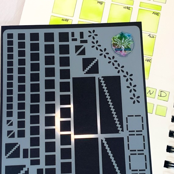 image shows close up of mylar stencil for layout basics showing boxes, squares, oblongs and dividers. poking out the top of the stencil is the central spiral of the open journal