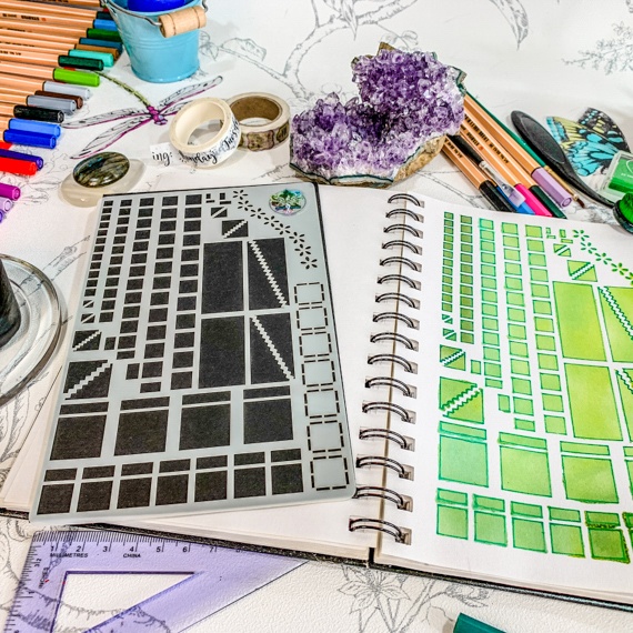 Image of the Journal Layouts Spreads Stencil, the stencil showing squares, oblongs and dividers is on a black background. the Stencil is lying on an open spiral journal, where the right had page is showing past the stencil where the stencil has been used with green inks and pens. above the journal are some brushes, inks and pens.