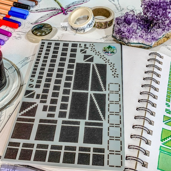 Image of the Journal Layouts Spreads Stencil, the stencil showing squares, oblongs and dividers is on a black background. the Stencil is lying on an open spiral journal, where the right had page is showing past the stencil where the stencil has been used with green inks and pens. above the journal are some brushes, inks and pens.