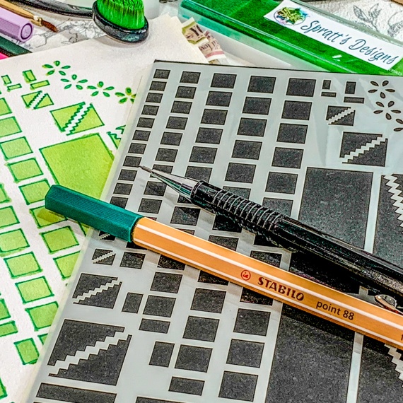 Image of the Journal Layouts Spreads Stencil, the stencil showing squares, oblongs and dividers is on a black background. the Stencil is lying on an open spiral journal, where the right had page is showing past the stencil where the stencil has been used with green inks and pens. above the journal are some brushes, inks and pens. Laying across the stencil is a propelling pencil and fine line green pen.