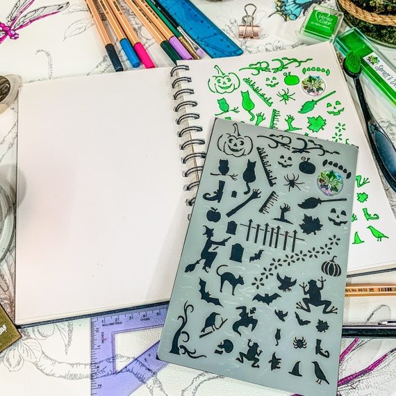 Magical Creatures Journal Stencil, Mythical wizardry Craft Stencil, Dragons, Fairies, witches, Pegasus, bats, cats, fairy, mermaids - The stencil is sitting on an open spiral bound journal, on the righthand page shows the stencil having been used with green inks, above the journal is fine line pens and below a triangle ruler, all on a pretty floral background.