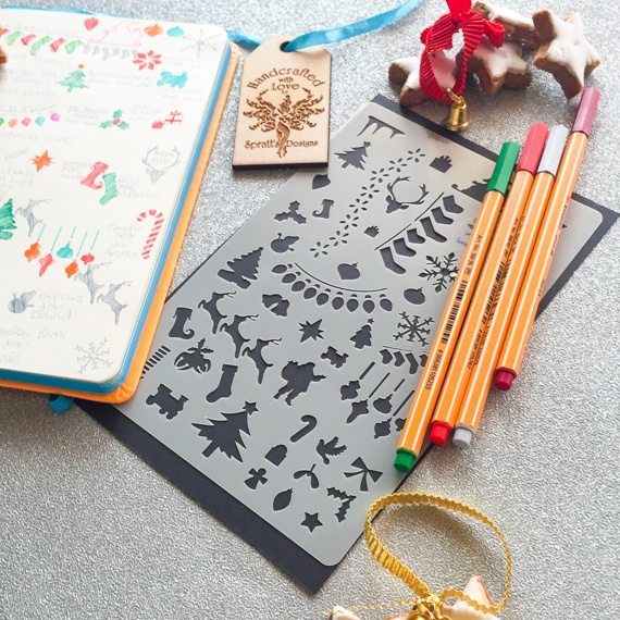 Christmas Journal Stencil laid on an open spiral journal, on a busy desk with pens and rulers around. The journal shows where the stencil has been used with coloured inks