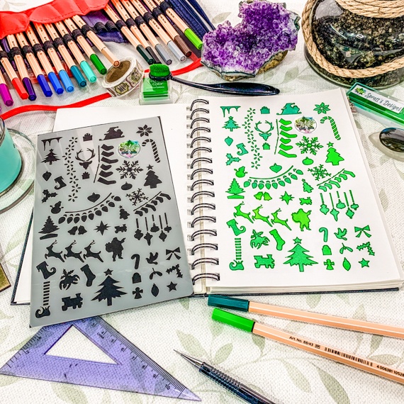 Christmas Journal Stencil laid on an open spiral journal, on a busy desk with pens and rulers around. The journal shows where the stencil has been used with green inks
