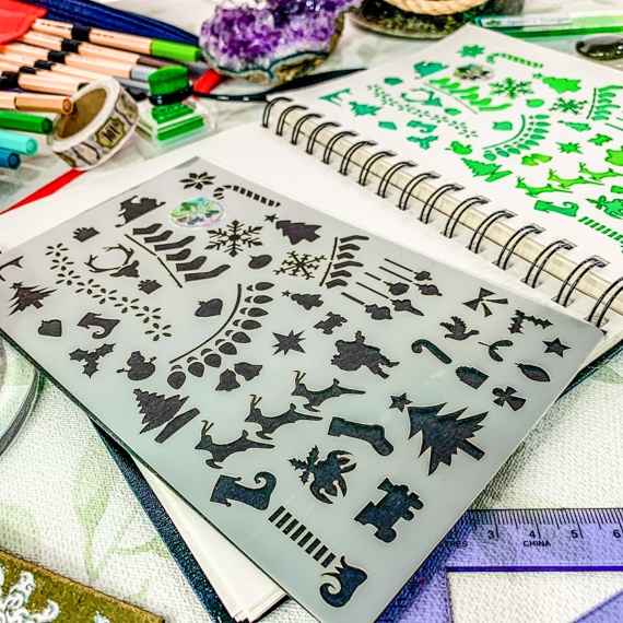 Christmas Journal Stencil laid on an open spiral journal, on a busy desk with pens and rulers around. The journal shows where the stencil has been used with green inks