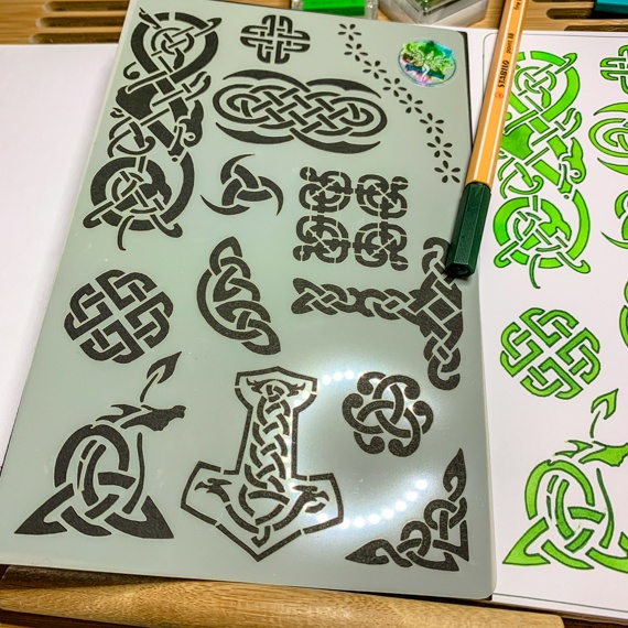 Celtic Journal Stencil Design 2 laying across an open journal where the stencil has been used with green inks and a pen laying across the page