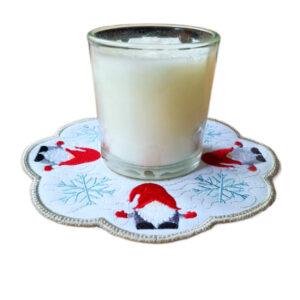 Small glass candle sitting on a festive embroidered coaster with a scalloped edge, featuring red gnomes and blue snowflake designs on a white background.