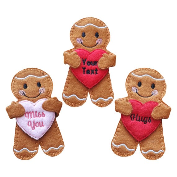 Three felt gingerbread characters holding colorful embroidered hearts. One heart is pink with the words 'Miss You,' another is red with 'Hugs,' and the middle red heart has space for custom text reading 'Your Text.' Each gingerbread figure has stitched details, smiling faces, and white icing accents.