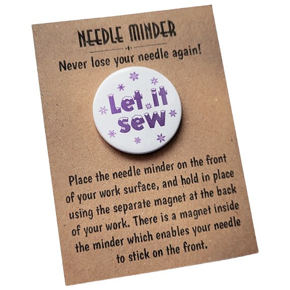 Round 38mm needle minder with the phrase “Let It Sew” in purple text surrounded by snowflakes, displayed on kraft backing card.