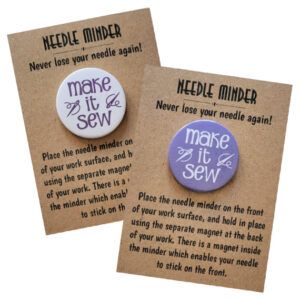 Two round needle minders, one with a white background and one with a purple background, both reading “Make It Sew” with scissor illustrations, displayed on kraft backing cards with instructions.