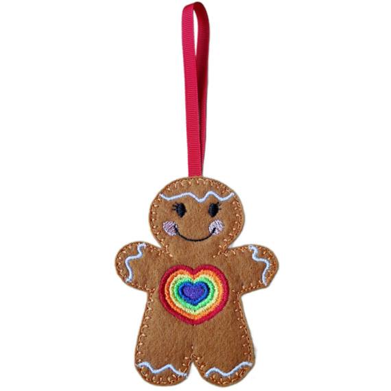 Rainbow Love Gingerbread Character – female version. A cheerful felt gingerbread decoration with delicate embroidered eyelashes, a vibrant rainbow heart on its front, smiling face, and white icing details, finished with a red ribbon loop for hanging.