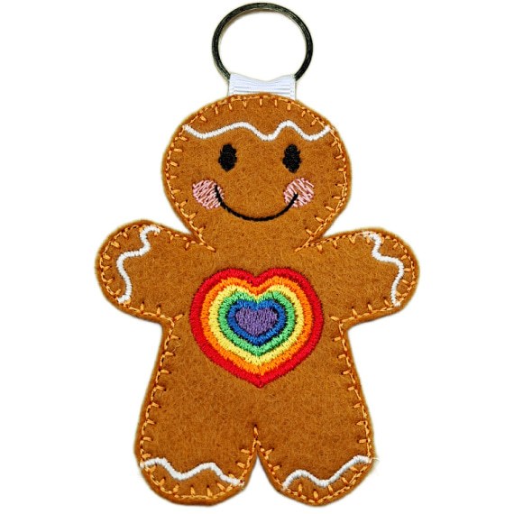 Rainbow Love Gingerbread Character – male version. A cheerful felt gingerbread keyring with a vibrant embroidered rainbow heart on its front, smiling face, and white icing details, finished with a silver keyring loop to carry with pride.