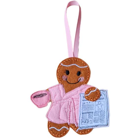 Rise and Shine Gingerbread Character — a handmade felt female gingerbread figure wearing a soft pink dressing gown with a matching belt, delicate embroidered eyelashes, and a pink ribbon loop for hanging. She is smiling while holding a pink cup of tea or coffee in one hand and a stitched newspaper in the other.