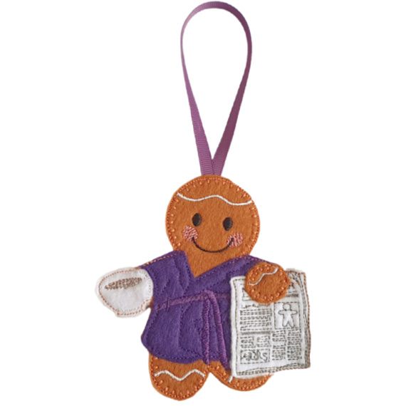 Rise and Shine Gingerbread Character — a handmade felt gingerbread figure wearing a purple dressing gown with a matching belt, holding a white cup of tea or coffee in one hand and a stitched newspaper in the other, with a purple ribbon loop for hanging.