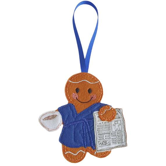 Rise and Shine Gingerbread Character — a handmade felt male gingerbread figure wearing a soft blue dressing gown with a matching belt, smiling while holding a white cup of tea or coffee in one hand and a stitched newspaper in the other. A blue ribbon loop is attached for hanging.