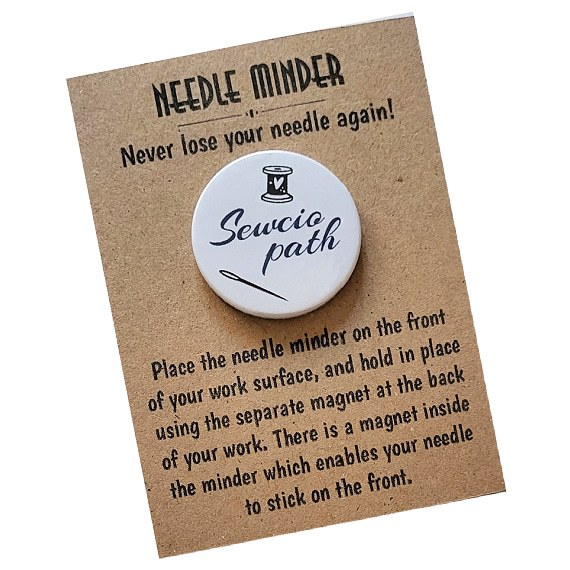 Round 38mm needle minder with the word “Sewciopath” in navy blue script, featuring a spool of thread and needle illustration, displayed on kraft backing card.