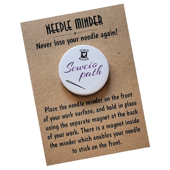 Round 38mm needle minder with the word “Sewciopath” in purple script, including spool of thread and needle design, displayed on kraft presentation card.