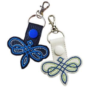 Celtic Dragonfly Keyring featuring intricate embroidery on faux leather, available in black with blue stitching and white with green stitching. Shaped like a dragonfly with Celtic knot details, it includes a snap button, keyring, and lobster clasp for easy attachment. Symbolises transformation, freedom, and new beginnings.