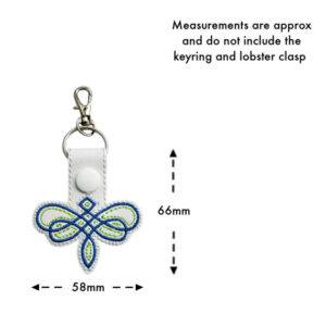 Celtic Dragonfly Keyring in white faux leather with green and blue embroidered Celtic knot design. Features a white snap button, keyring, and lobster clasp. Measurements shown: approximately 66mm in height and 58mm in width (excluding keyring and clasp). Text indicates measurements are approximate.
