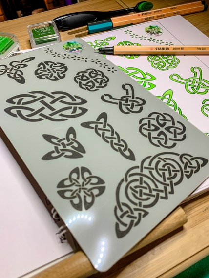 Celtic Knots Design 3 Journal Stencil - The stencil with multiple Celtic knots lays on an open journal where the right hand page shows the stencils shapes laid out in green inks, there is a pen laying across the page.