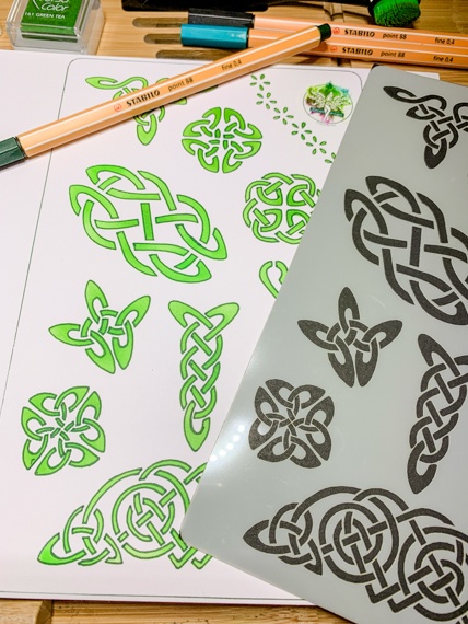 Celtic Knots Design 3 Journal Stencil - A close up of the left hand edge of the stencil showing a few knots and behind a page of a journal showing the knots in green inks. A pen lays across the page.