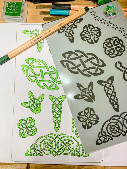 Celtic Knots Design 3 Journal Stencil - A close up of the left hand edge of the stencil showing a few knots and behind a page of a journal showing the knots in green inks. A pen lays across the page.