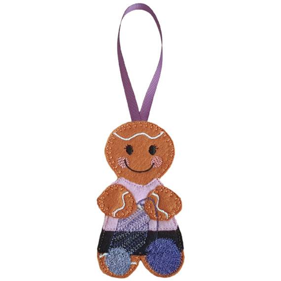 A felt crochet gingerbread character hanging decoration with a purple ribbon loop, featuring a smiling face, cosy stitched details, a pink top, black skirt, and balls of yarn.