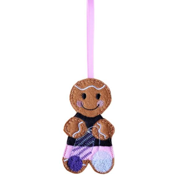A felt crochet gingerbread character hanging decoration with a pink ribbon loop, featuring a smiling face, cosy stitched details, a dark top, black skirt, and balls of yarn.