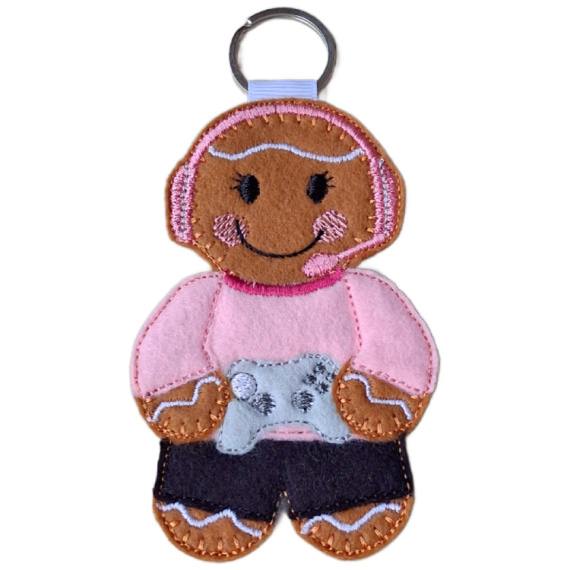 A felt female gamer gingerbread keyring with a silver ring, wearing a pink top, black trousers, and a pink gaming headset, smiling while holding a light grey game controller.