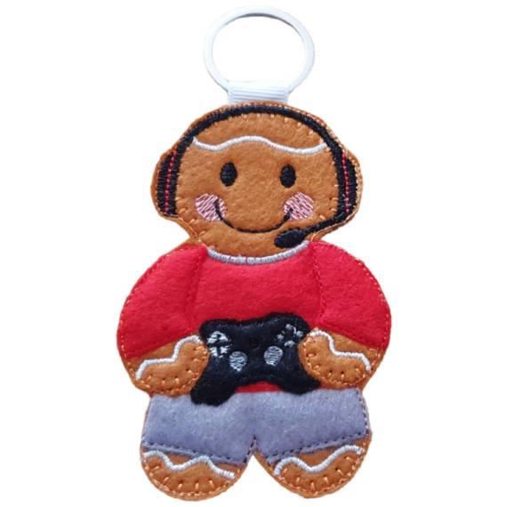 A felt male gamer gingerbread keyring with a silver ring, wearing a red top, grey trousers, and a black gaming headset, smiling while holding a black game controller.