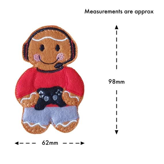 A felt gamer gingerbread character wearing a red top, grey trousers, and a black gaming headset, holding a small black game controller. The character measures approximately 98 mm tall and 62 mm wide.