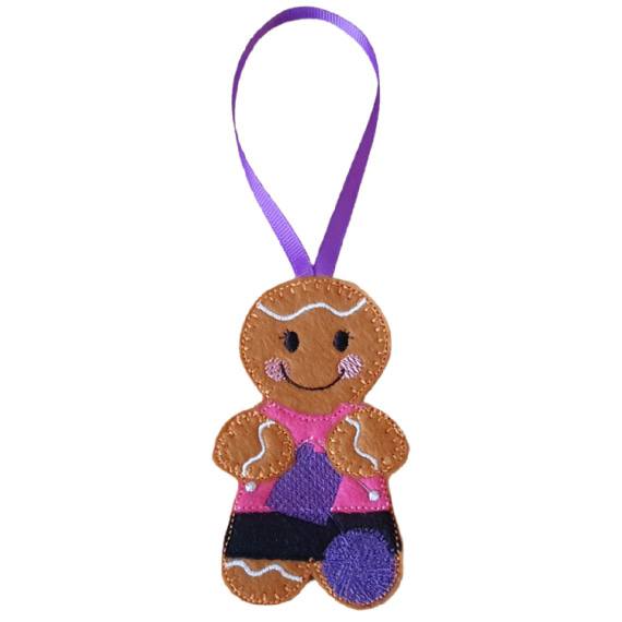 A felt knitting gingerbread character hanging decoration with a purple ribbon loop, featuring a smiling face, cosy stitched details, a pink top, patterned skirt, and a ball of yarn.