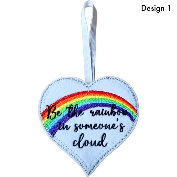 Heart-shaped felt hanging decoration labelled Design 1, featuring an embroidered rainbow and the message ‘Be a rainbow in someone’s cloud’, with a ribbon loop at the top.
