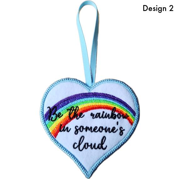 Heart-shaped felt hanging decoration labelled Design 2, featuring an embroidered rainbow and the message ‘Be a rainbow in someone’s cloud’, with a ribbon loop at the top.