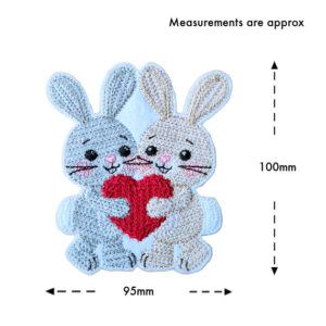 Two felt bunnies hugging and holding a red heart, machine embroidered with a knit stitch pattern, shown on a white background with approximate measurements of 95mm wide and 100mm tall.