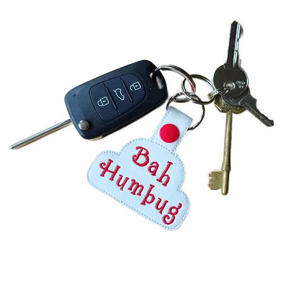 A set of keys on a keyring, featuring a white faux leather keyring embroidered with the words ‘Bah Humbug’ in red lettering and a red snap fastener, attached alongside a car key fob and two metal keys.