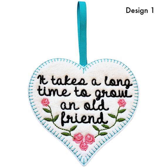 Heart-shaped felt hanging decoration labelled Design 1, featuring an embroidered friendship message reading, ‘It takes a long time to grow an old friend’, with floral details and a ribbon loop at the top.