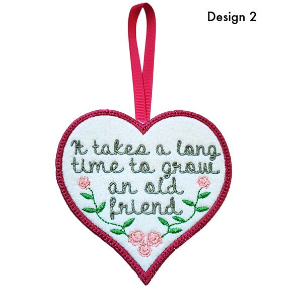 Heart-shaped felt hanging decoration labelled Design 2, featuring an embroidered friendship message reading, ‘It takes a long time to grow an old friend’, with floral details and a ribbon loop at the top.