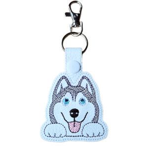 A cute Kawaii Husky keyring made from white faux leather, featuring an embroidered husky face with blue eyes, a smiling expression, and a pink tongue, attached to a silver keyring with a light grey snap fastener and lobster clasp.