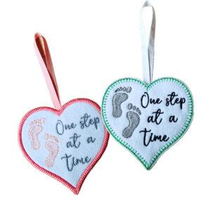 Two embroidered felt heart hanging decorations with footprints and the words 'One Step at a Time', one with a pink border and ribbon and one with a green border and white ribbon.