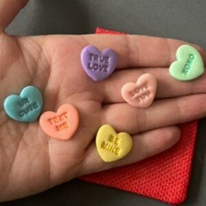 Hand holding six handmade polymer clay pastel love heart tokens with embossed sparkly phrases, inspired by love heart sweets, shown with a red drawstring gift pouch