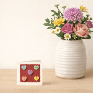 Small handmade greeting card with a cream background and torn red sugar paper centre, decorated with five colourful polymer clay heart grounding stones, photographed beside a white vase of flowers on a wooden surface.