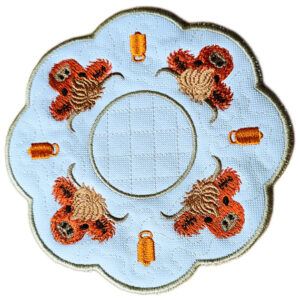 Close-up of a round scalloped candle mat in white fabric, embroidered with four Highland cow faces arranged in a circle around a stitched centre, with small cow bell motifs between each cow.