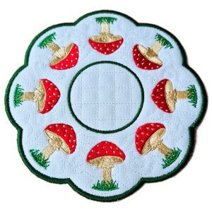 Close-up of a round scalloped candle mat embroidered with toadstools arranged in a circular pattern around a stitched centre, finished with a contrasting stitched border.